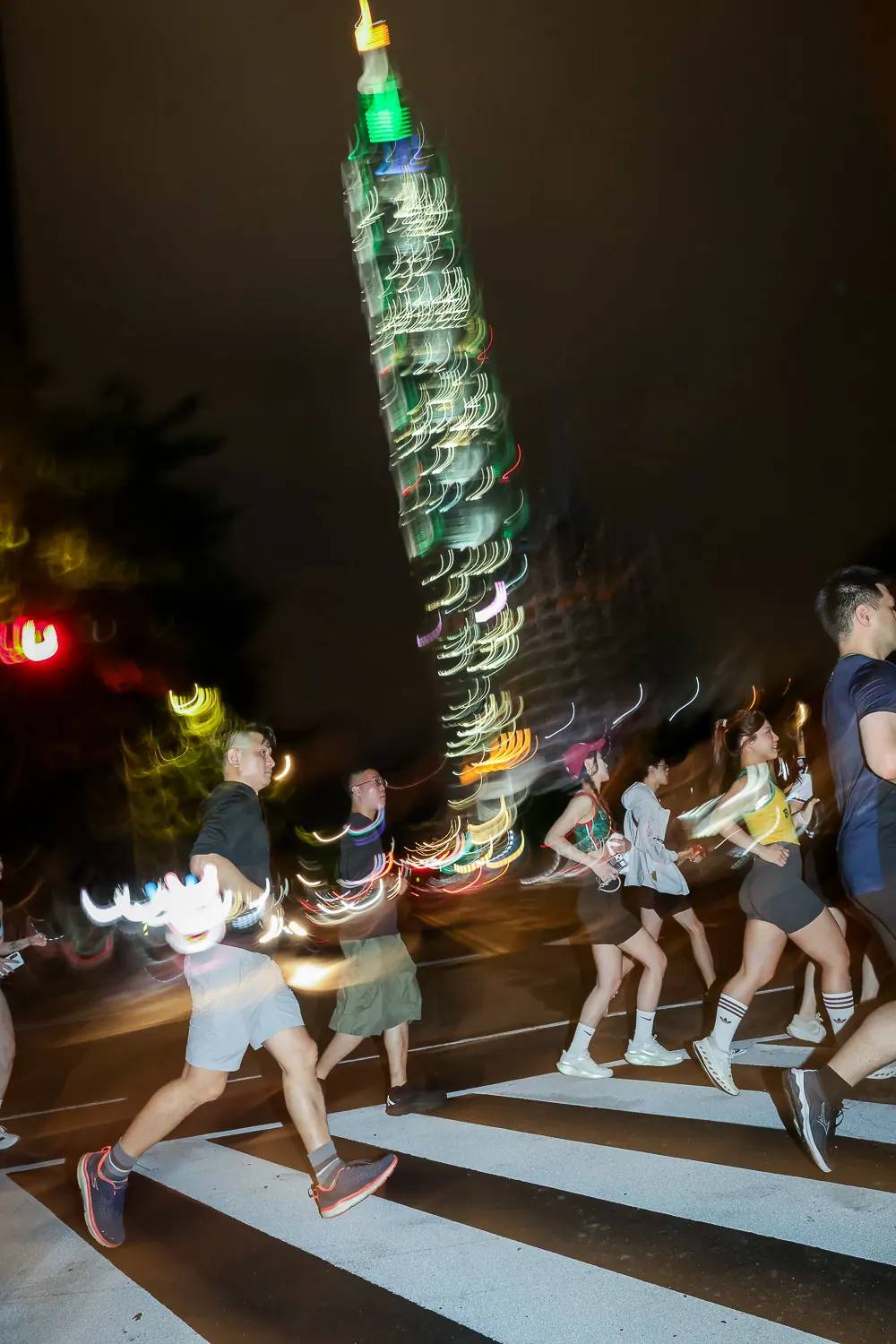 Runners crossing a street at night with Taipei 101 lit up behind them, blurred by motion.